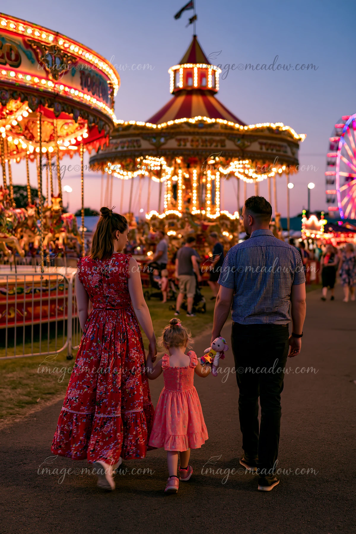 A family enjoys a fairground carousel while walking through a amusement park at dusk.