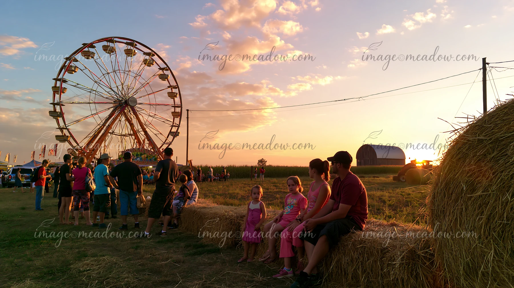 A family is seated on hay bales while waiting for the ferris wheel at a fair during sunset.