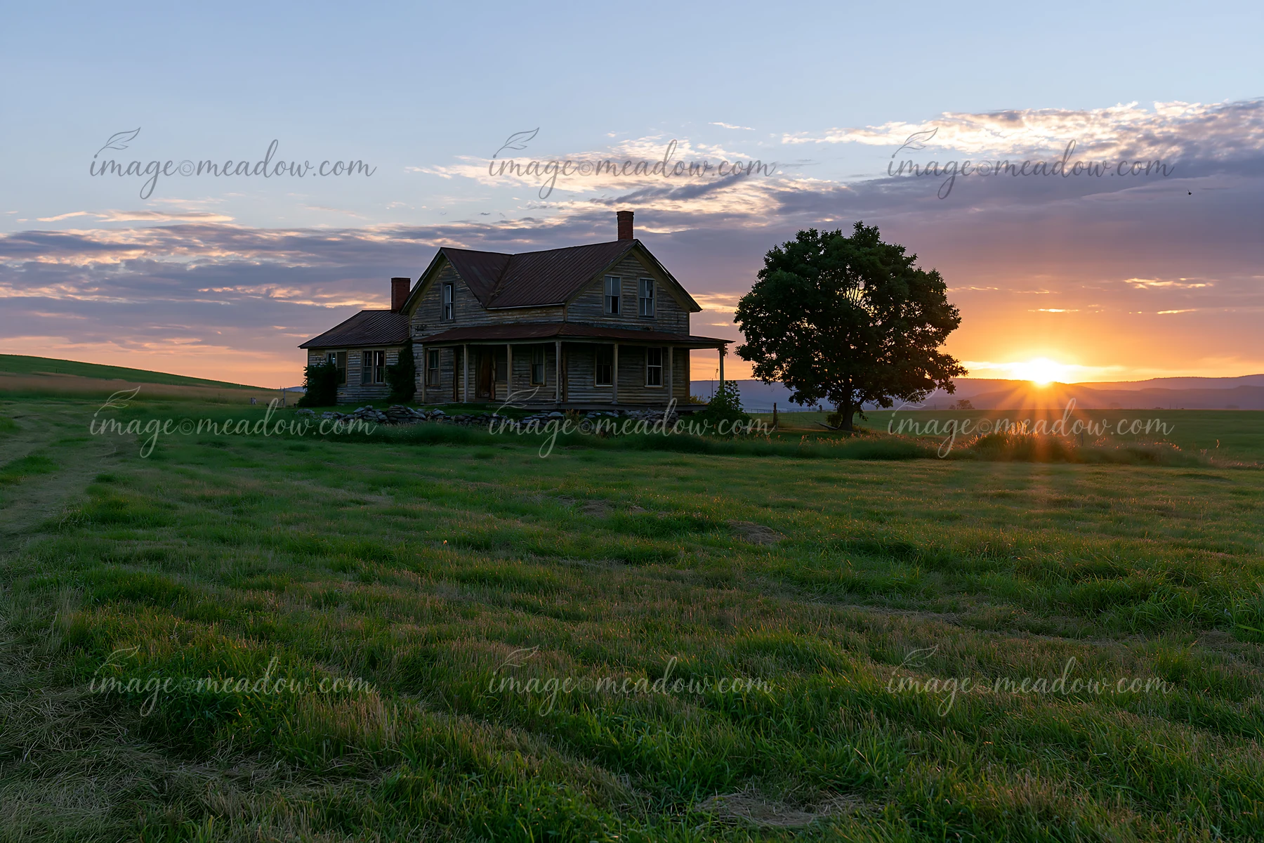 A farmhouse sits in a green field as the sun sets in the background, creating a warm glow in the sky.