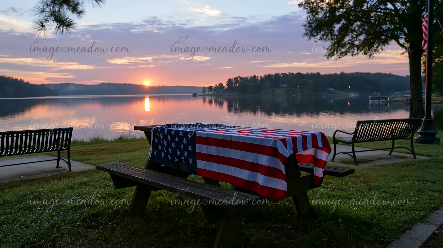 A flag is draped over a picnic table near a lake as the sun rises.