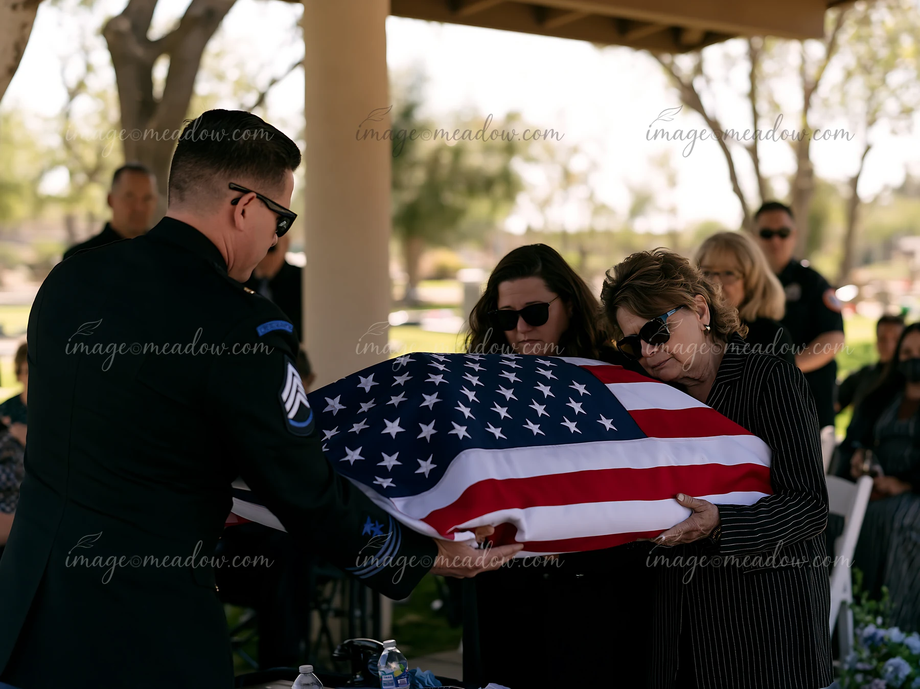 A folded American flag presented to a grieving family at a funeral service, with mourners seated in the background under a shaded area.