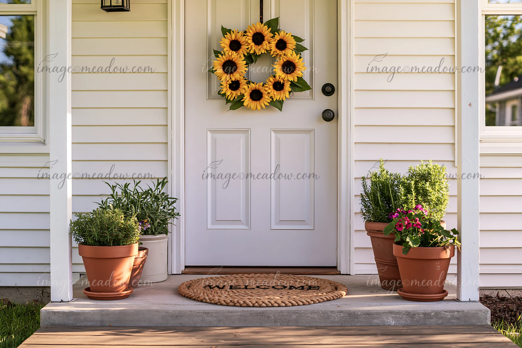A front door with wreath and welcome doormat is decorated with sunflowers and surrounded by potted plants.