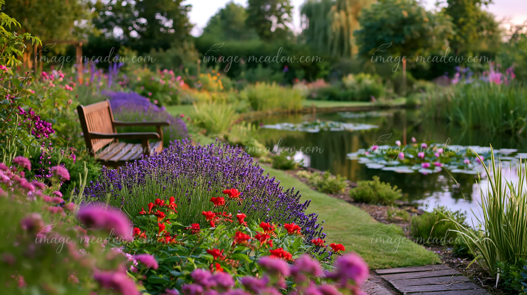 A garden bench sits beside a water lily pond surrounded by vibrant flowers and greenery.