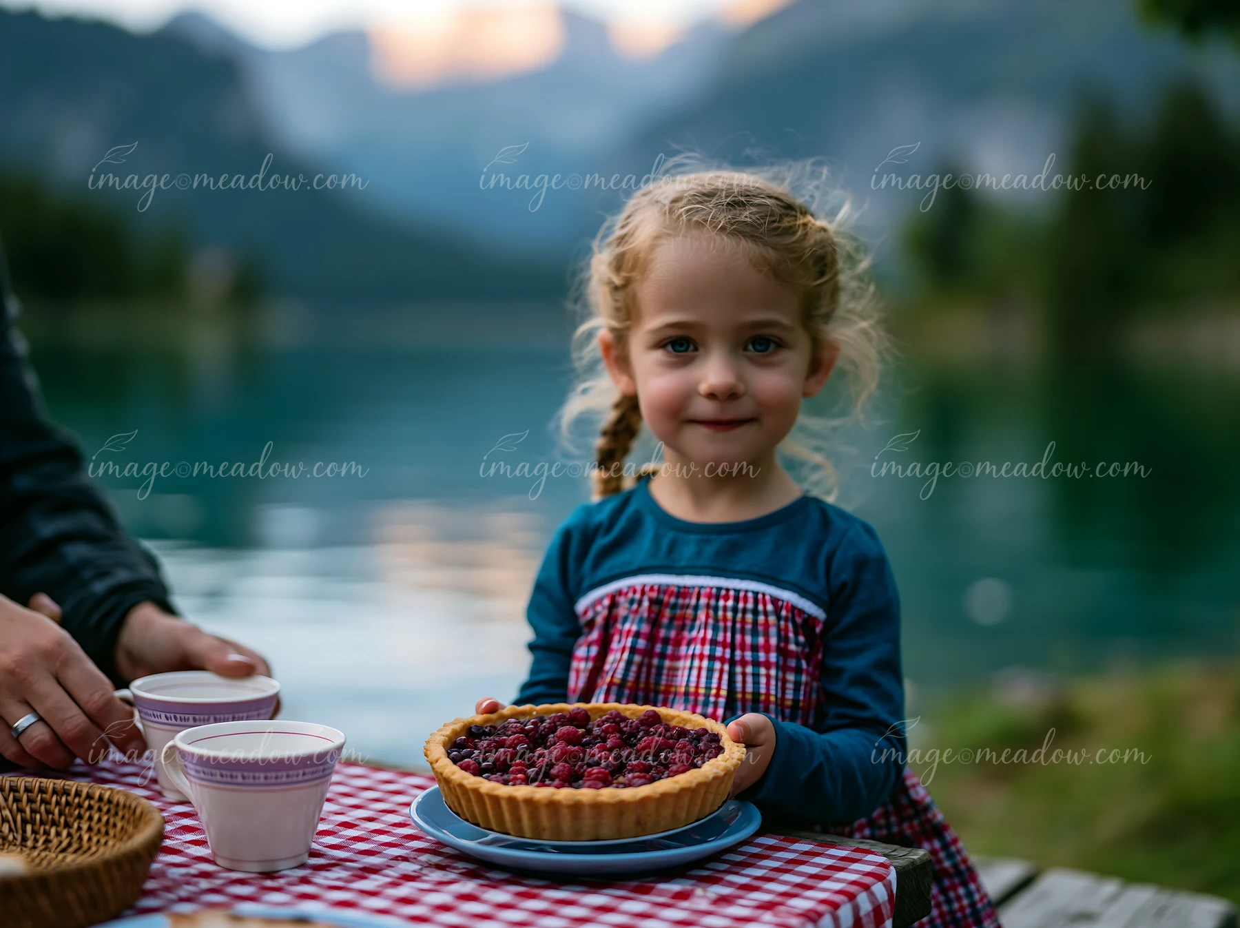 A girl holds a berry tart while sitting by an alpine lake with mountains in the background.