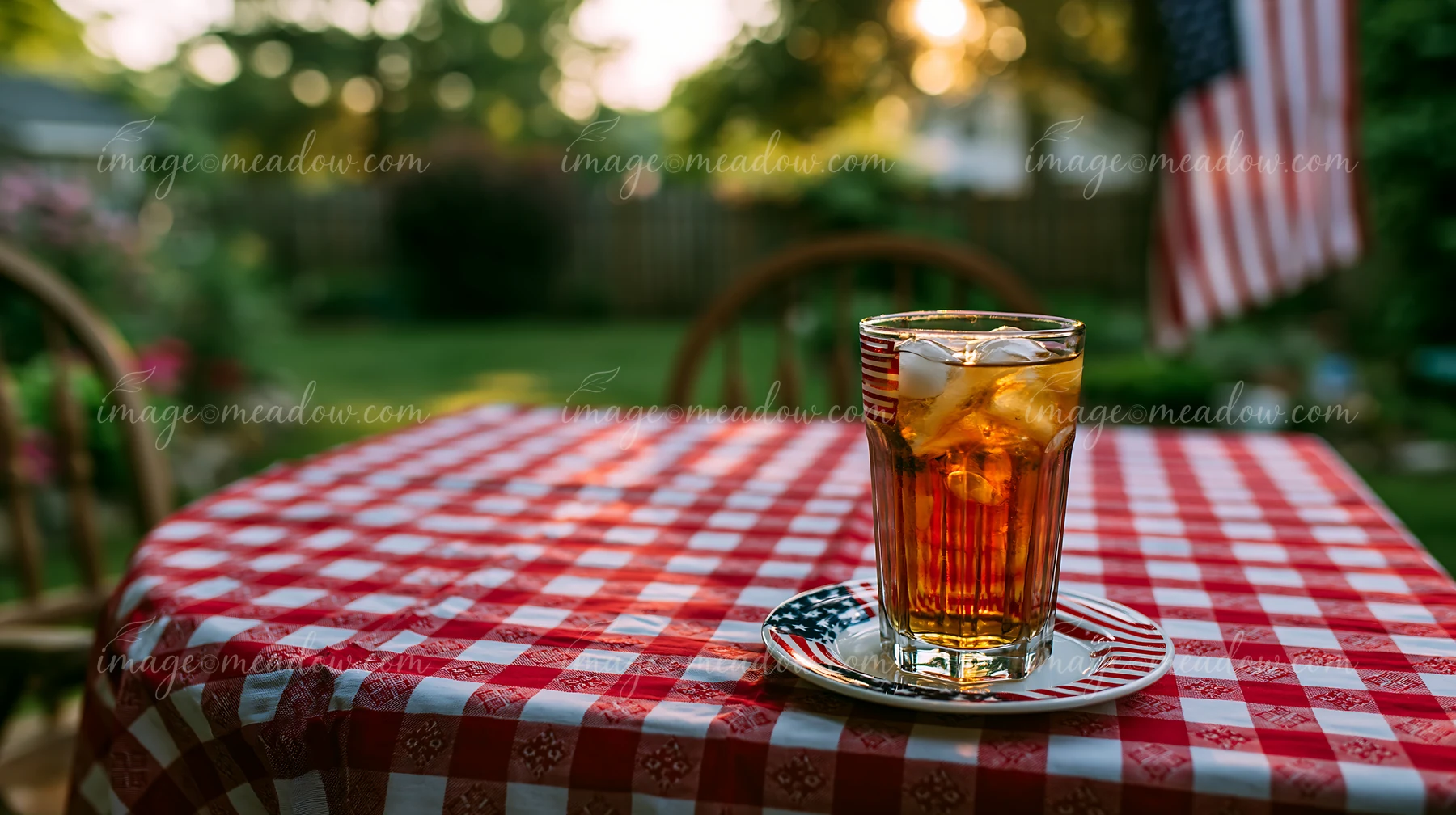 A glass of iced tea sits on a plate on a red and white checkered tablecloth outdoors.