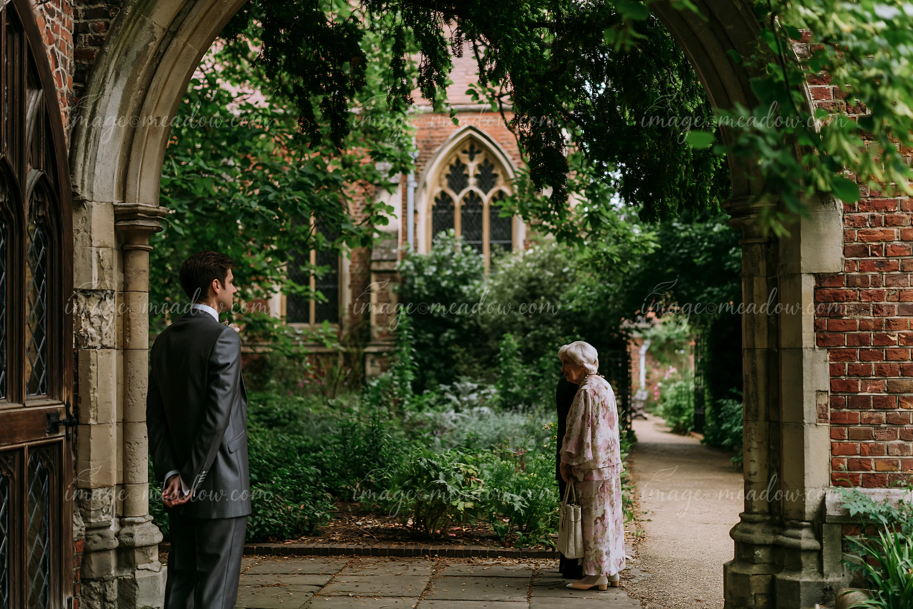 A groom stands near a church arch, waiting while an elderly woman approaches.