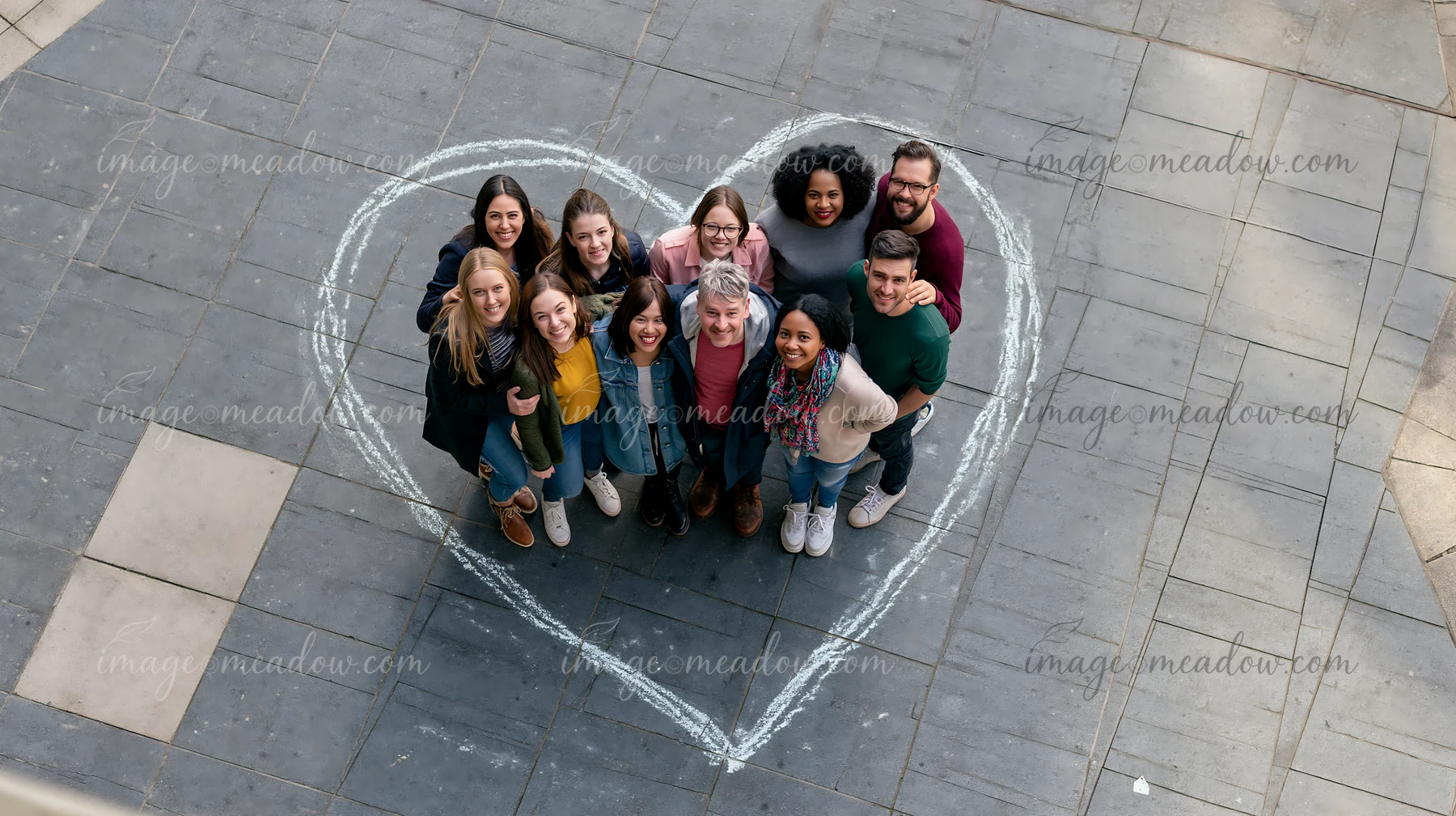A group of diverse people stands together inside a heart-shaped chalk outline on pavement in an outdoor setting.