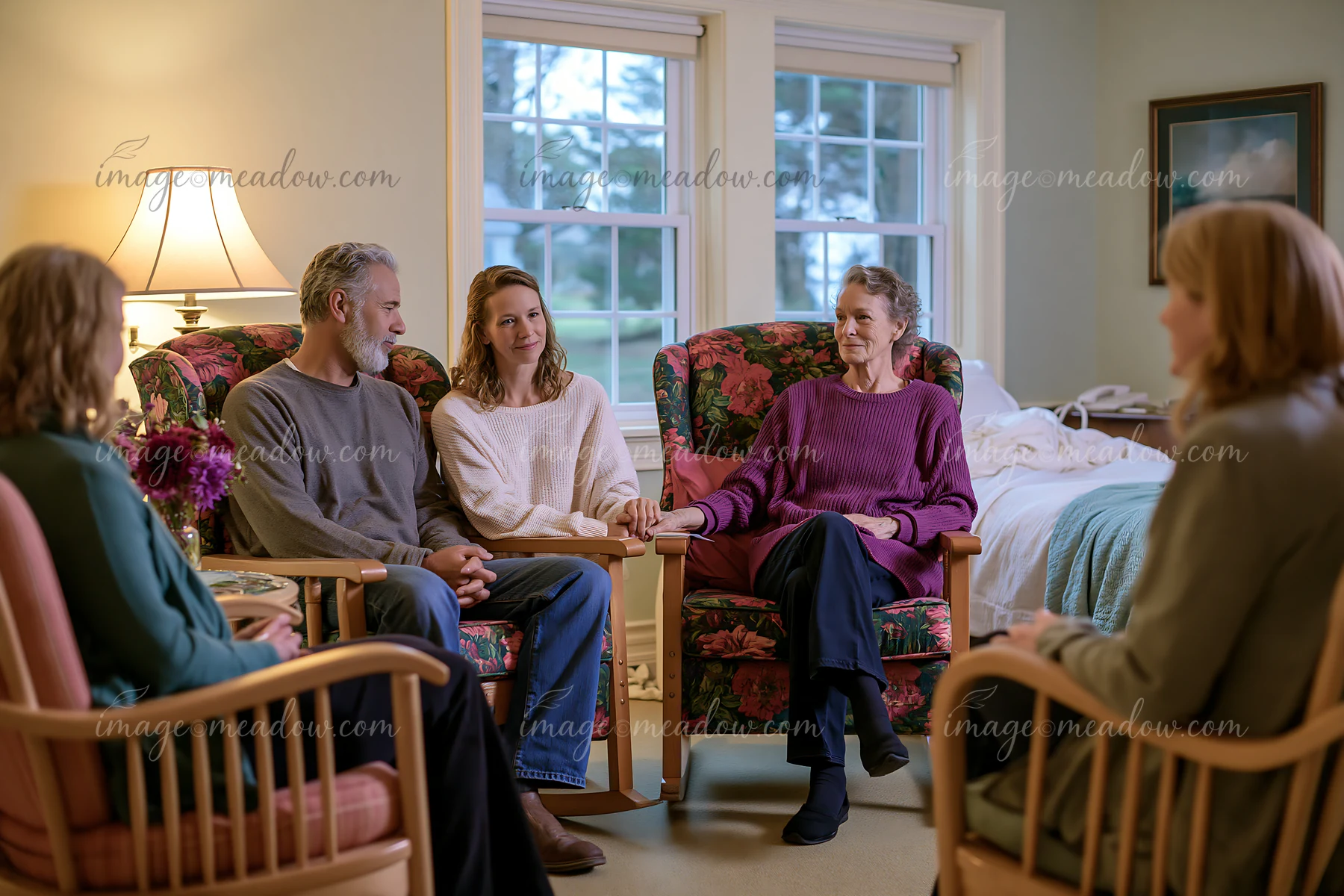 A group of five people sit in a hospice room, holding hands together while supporting a patient.