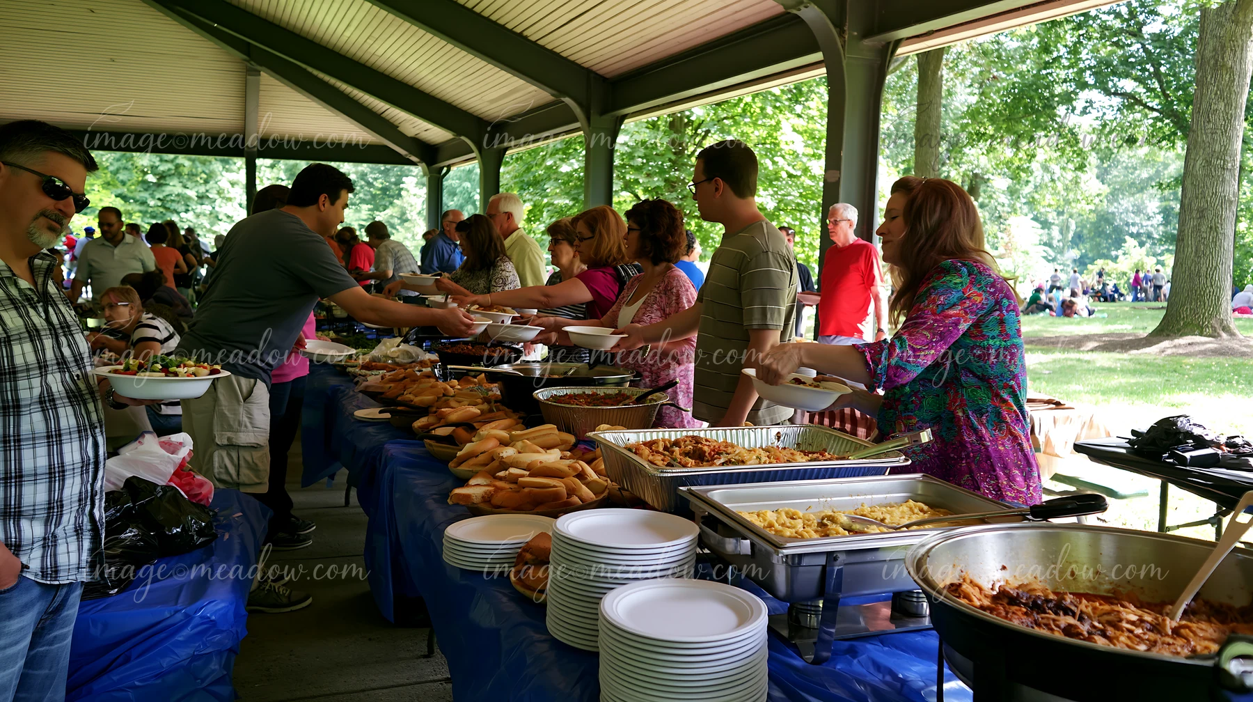 A group of people gather around a buffet table at a church event in a park, serving food onto their plates as others socialize nearby.