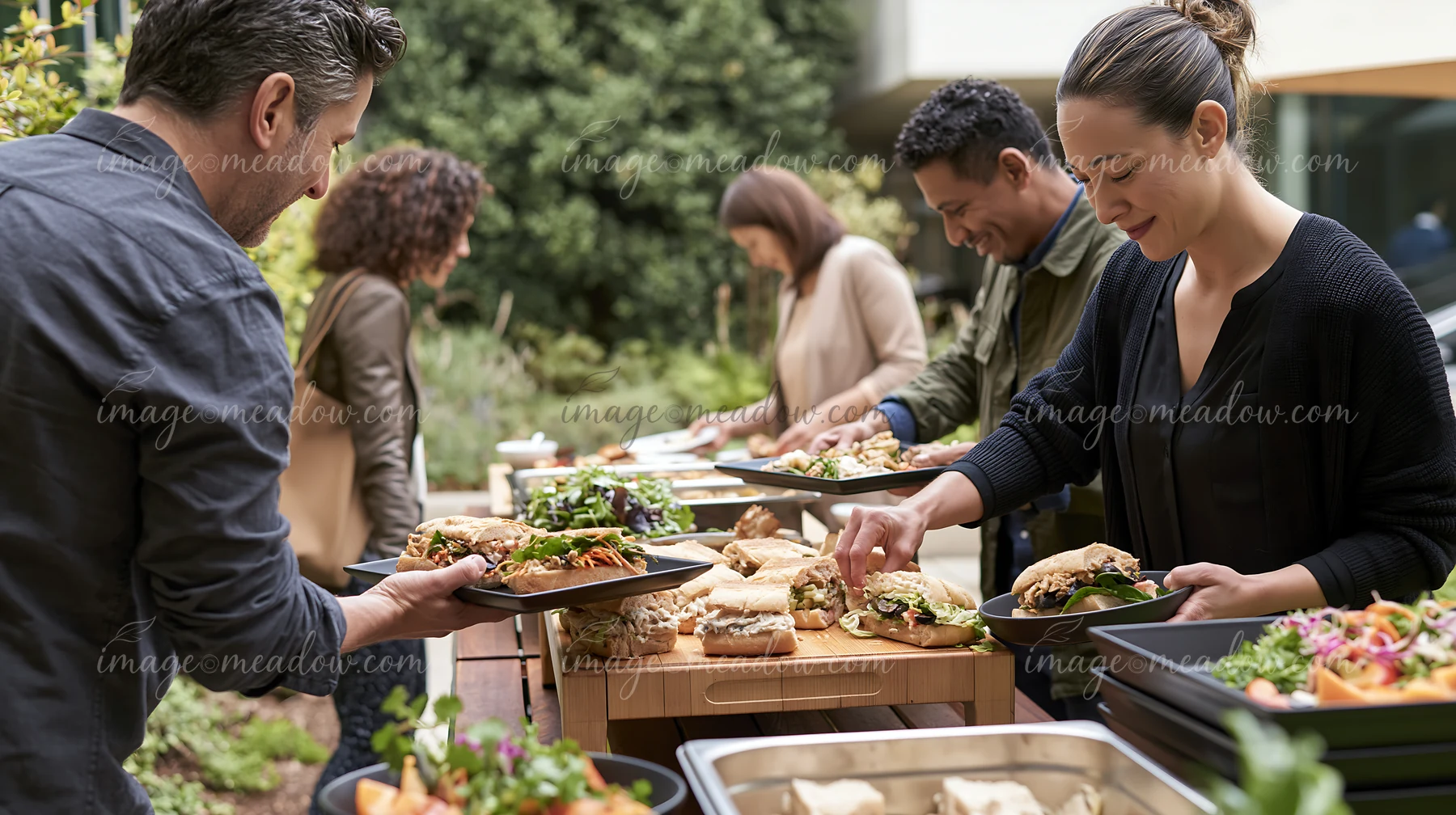 A group of people is enjoying a sandwich buffet for workplace lunch outdoors, helping themselves to a variety of sandwiches and salads at a long table in a gard