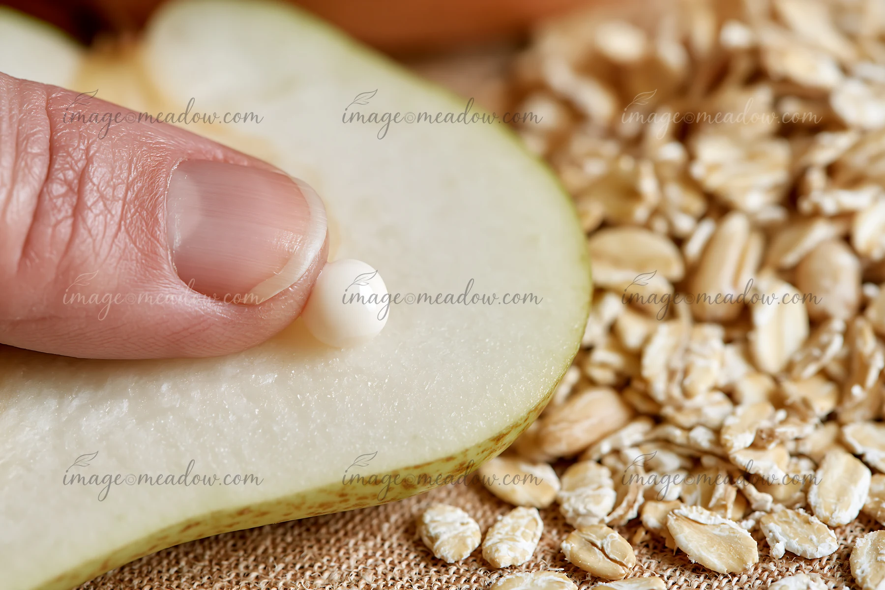 A hand applies a small cream dose with natural skincare ingredients to a slice of pear next to oats on a textured surface.