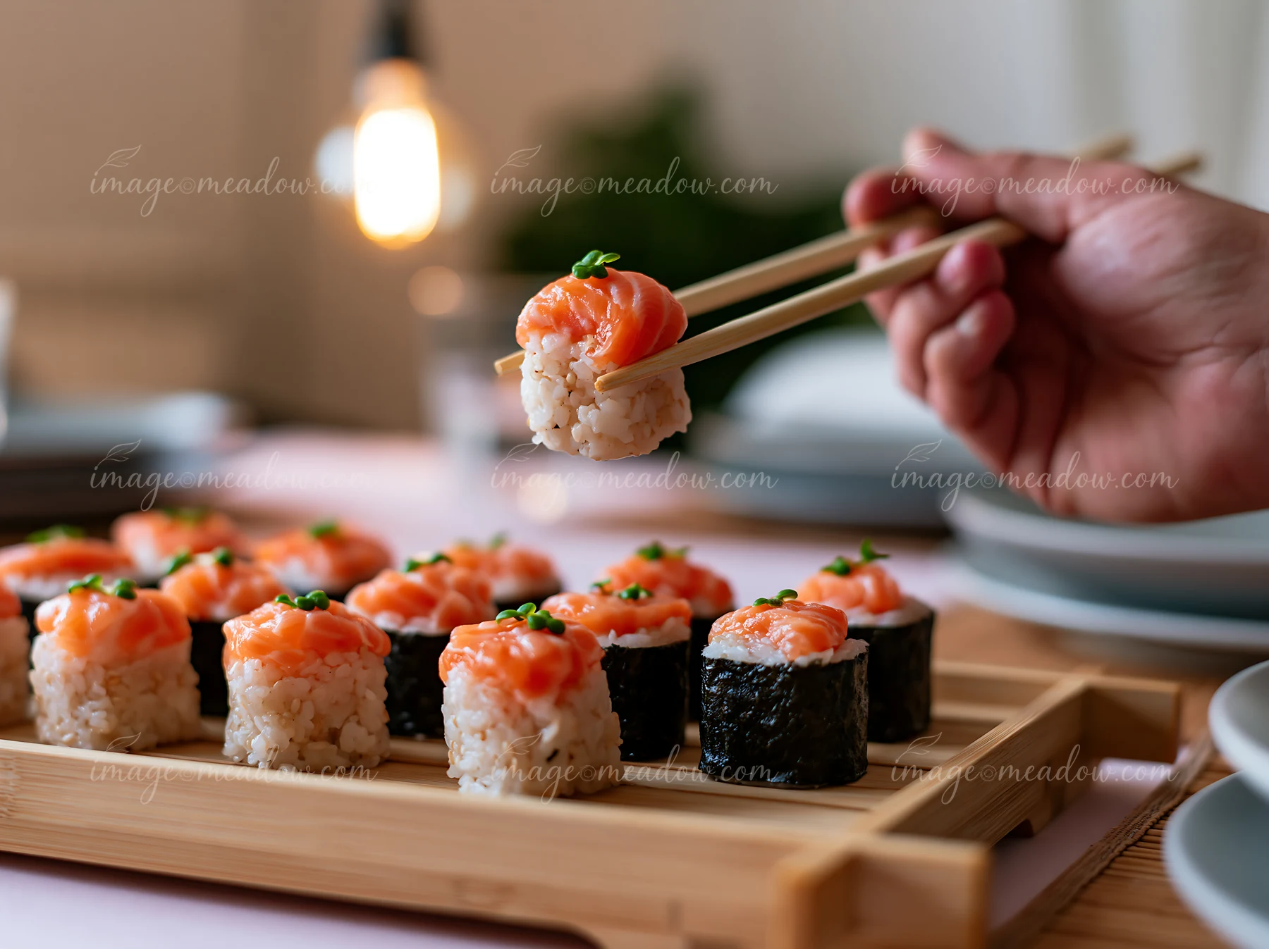 A hand holds a single sushi piece ready to eat with chopsticks, surrounded by other sushi pieces on a wooden tray.