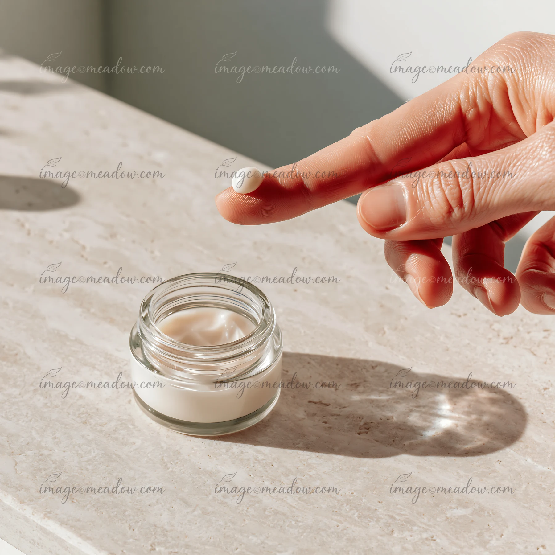 A hand holds a small amount of cream over a glass jar placed on a light colored surface.