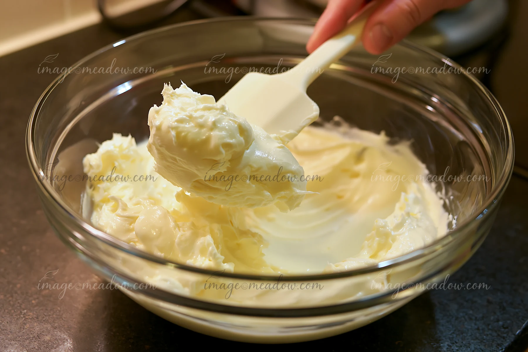 A hand holds a spatula as it lifts a thick cream dose with smooth glossy texture from a glass mixing bowl on a dark countertop, showing the creamy mixture ready