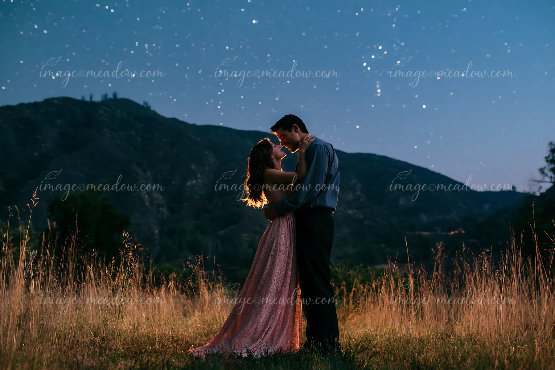 A happy couple kissing beneath stars in nature during the night with mountains in the background.