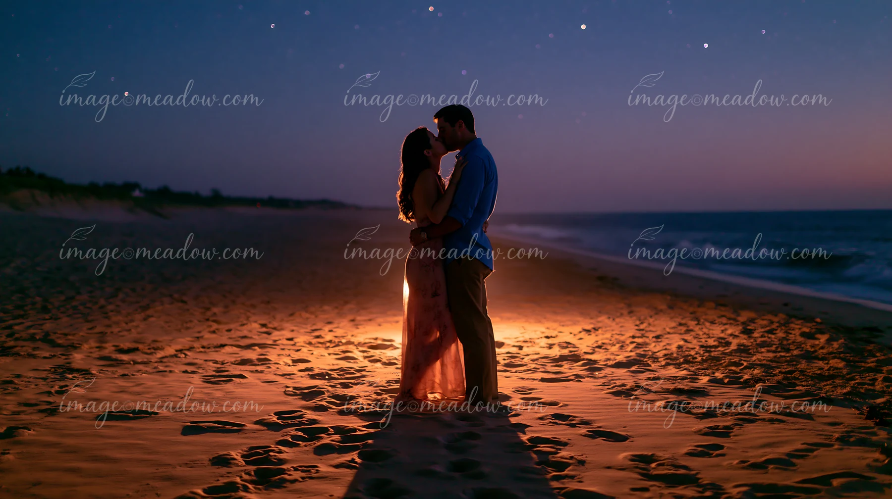 A happy couple kissing beneath stars in nature stands on a beach at dusk.