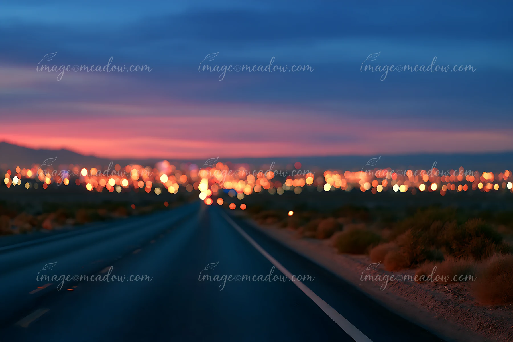 A highway stretches towards a city in the distance as dusk falls, with bright lights illuminating the urban landscape.