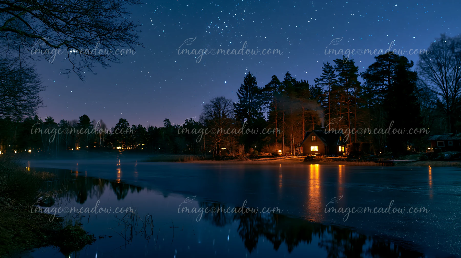 A house emits smoke from its chimney, illuminated by starlight, near a still lake.