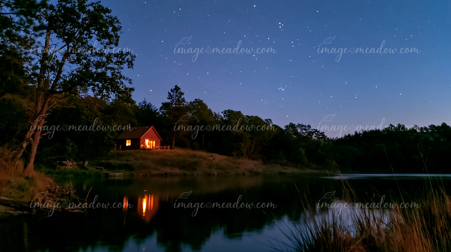 A house on a hill beneath stars shines brightly at night, reflecting on calm water.