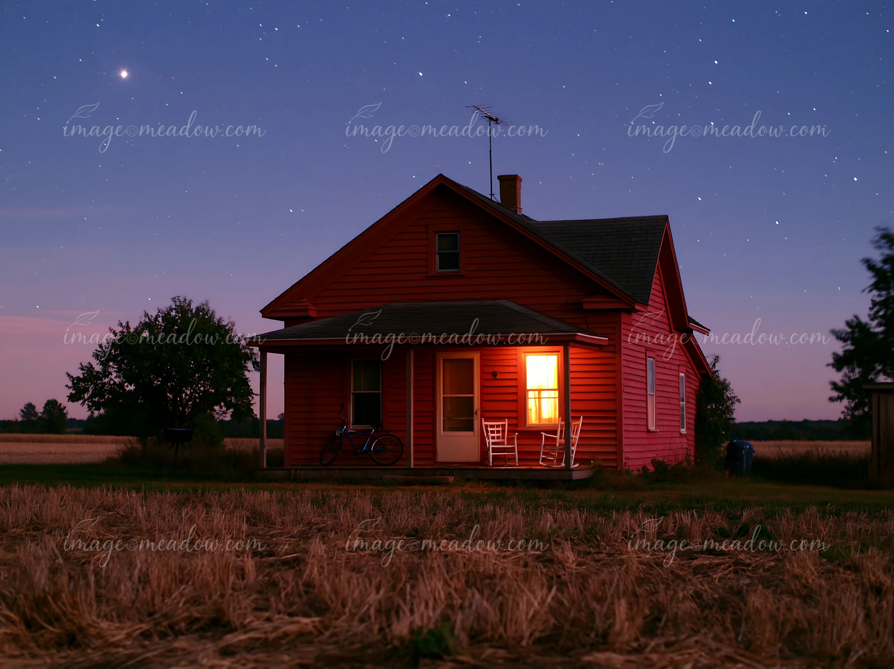 A house stands in a field with a bright window illuminated against a night sky full of stars.