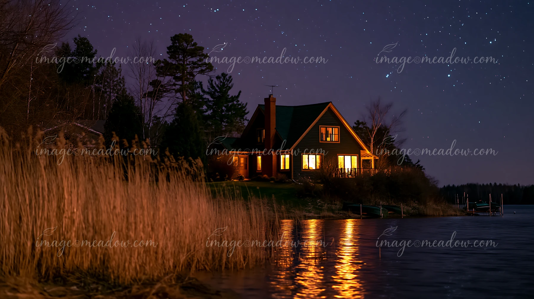 A house under a starry sky with warm lights sits by a lake, with reflections on the water.