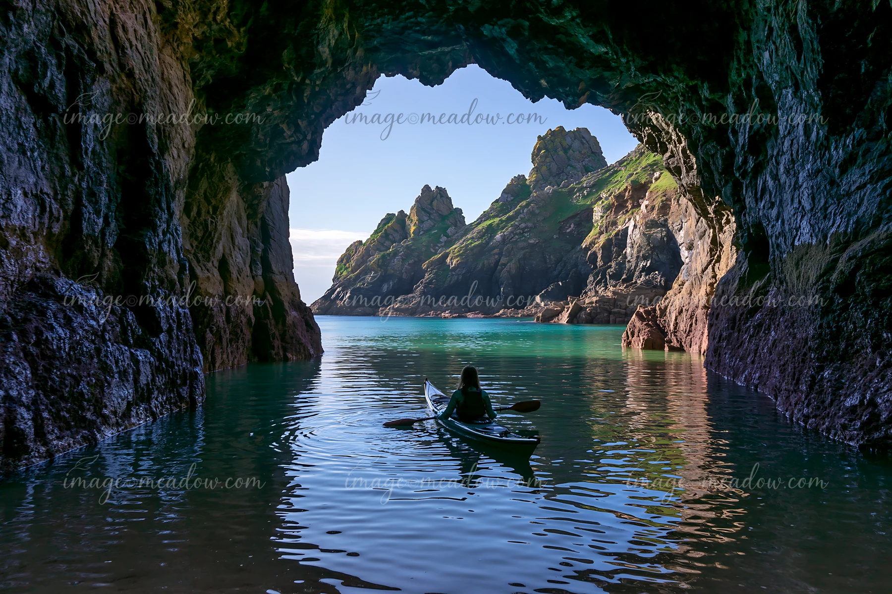 A kayaker paddles through a sea cave, with a clear view of a rocky coastline outside.