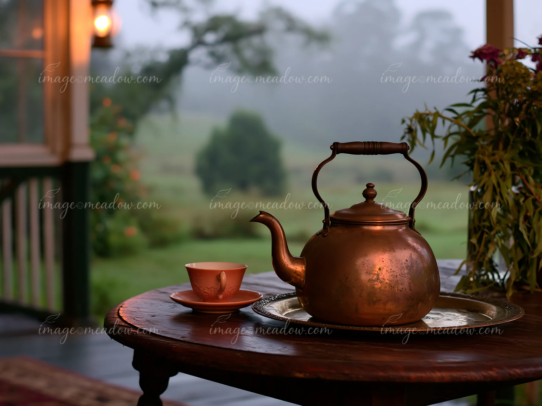 A kettle sits on a table beside a cup in a misty countryside setting.