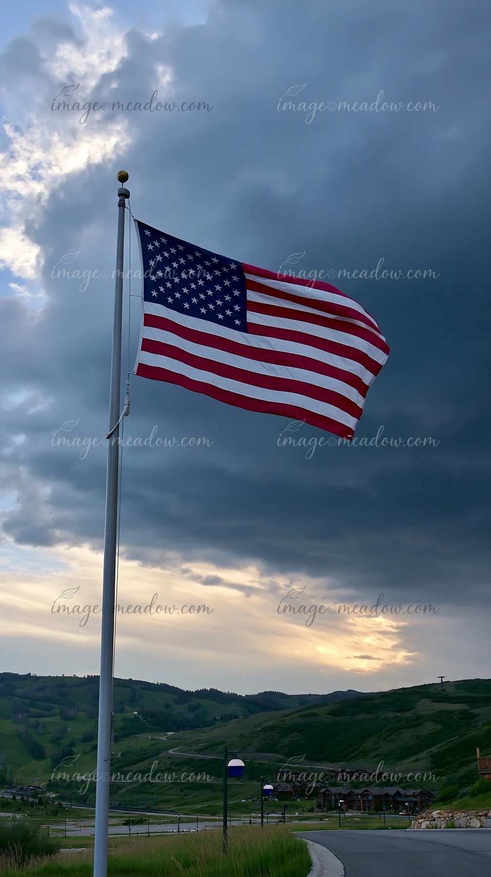 A large American flag is flying in strong wind on a pole, set against a backdrop of dramatic clouds.