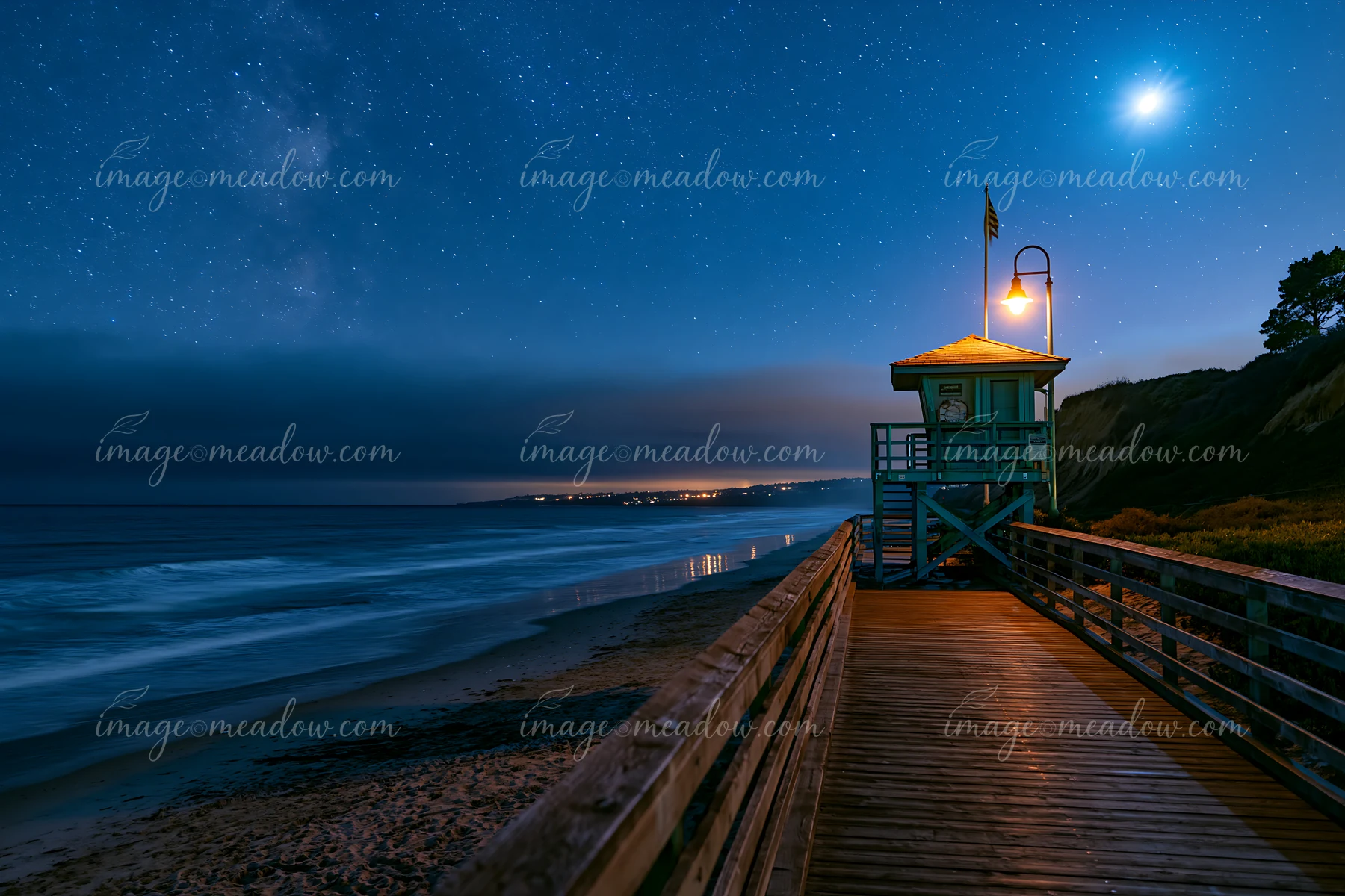 A lifeguard tower stands at the end of a coastal boardwalk under a starry night sky.