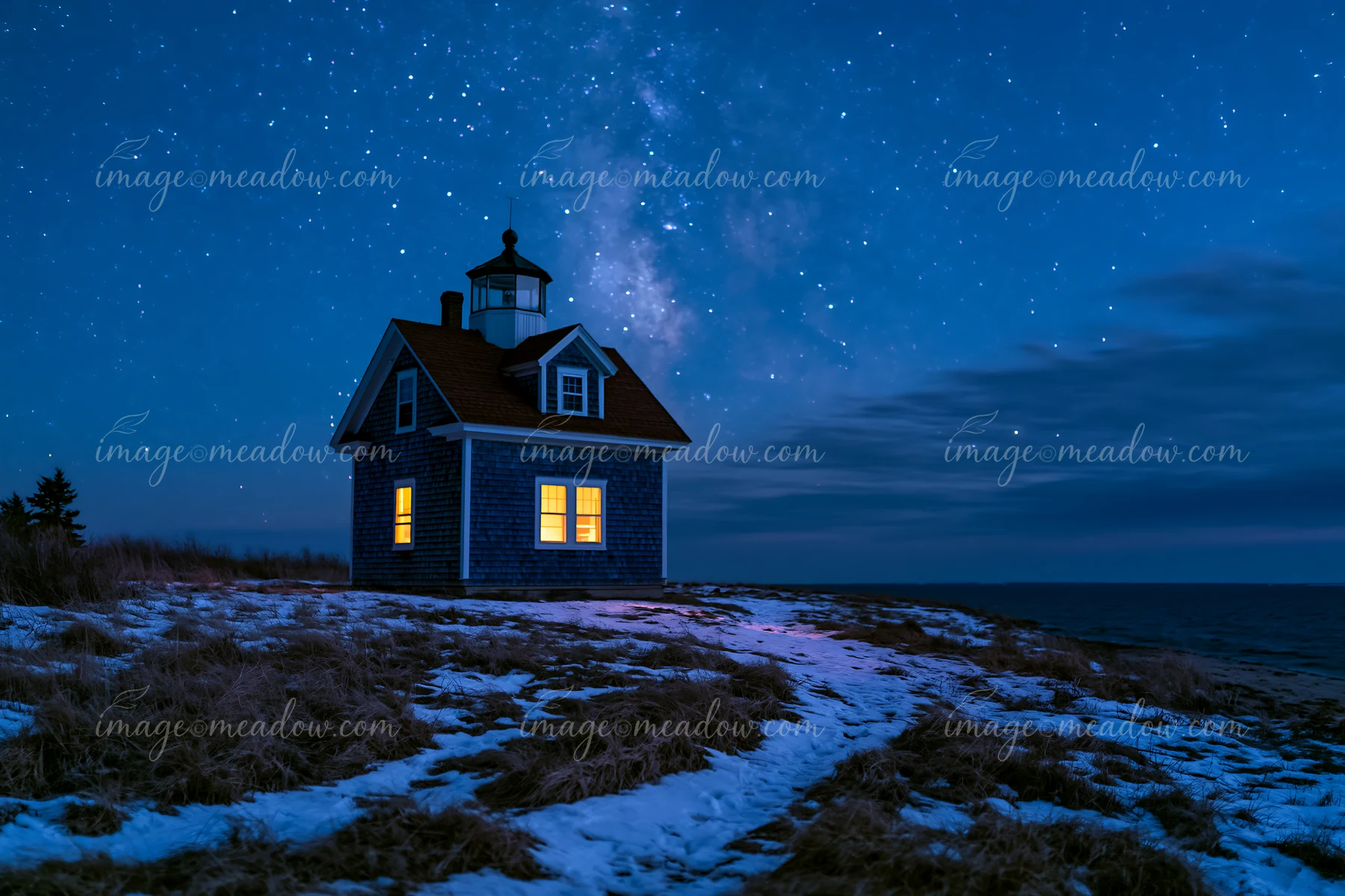 A lighthouse house stands on a grassy area near the shore under a starry sky.