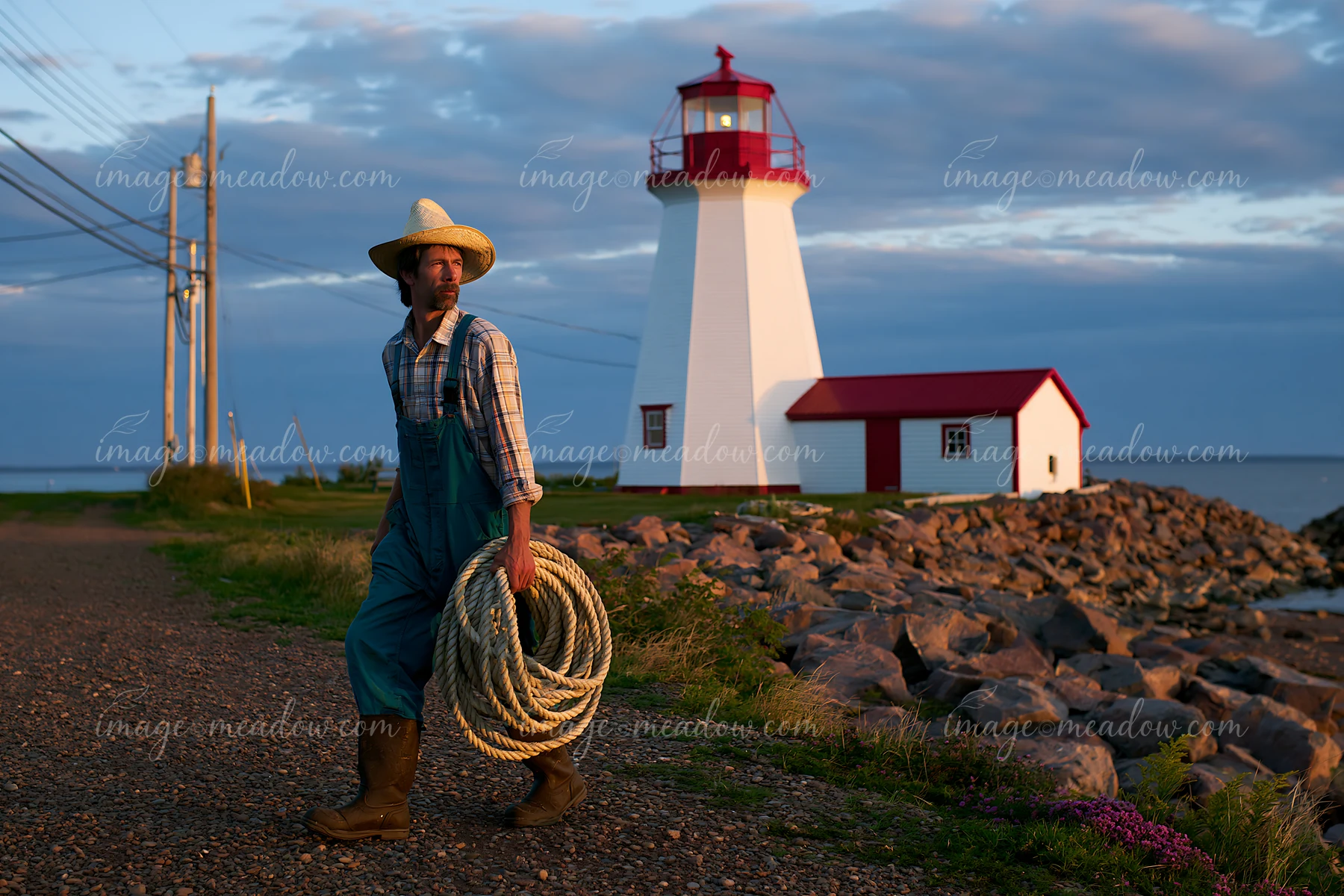A lighthouse maintenance worker at work walks along the path near a coastal lighthouse, carrying rope.