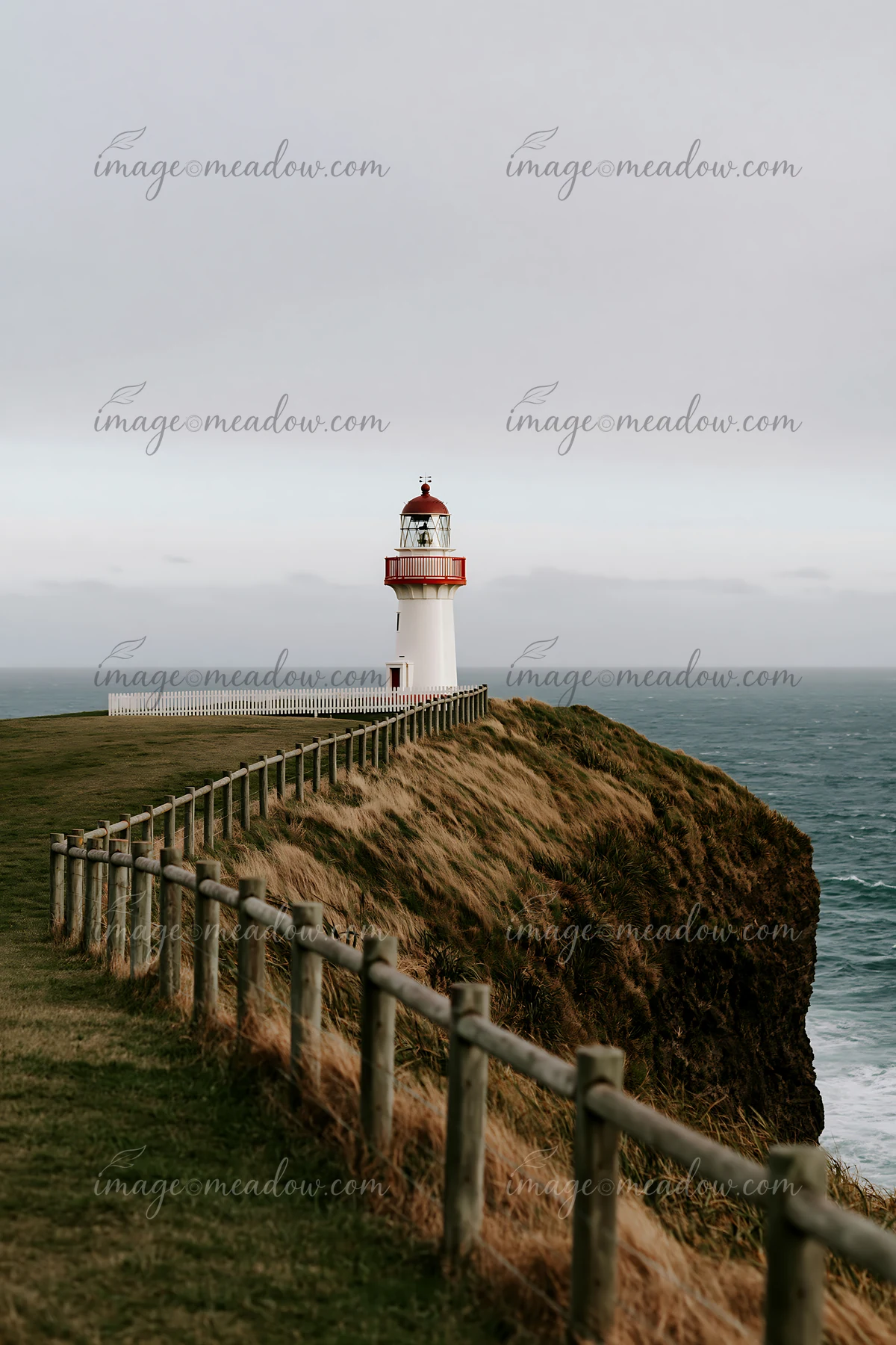 A lighthouse stands on a cliff overlooking the ocean, surrounded by grass and a wooden fence.