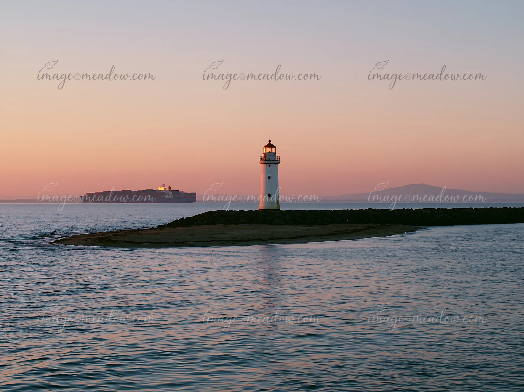 A lighthouse stands on a rocky point as a cargo ship travels in the water at sunset.