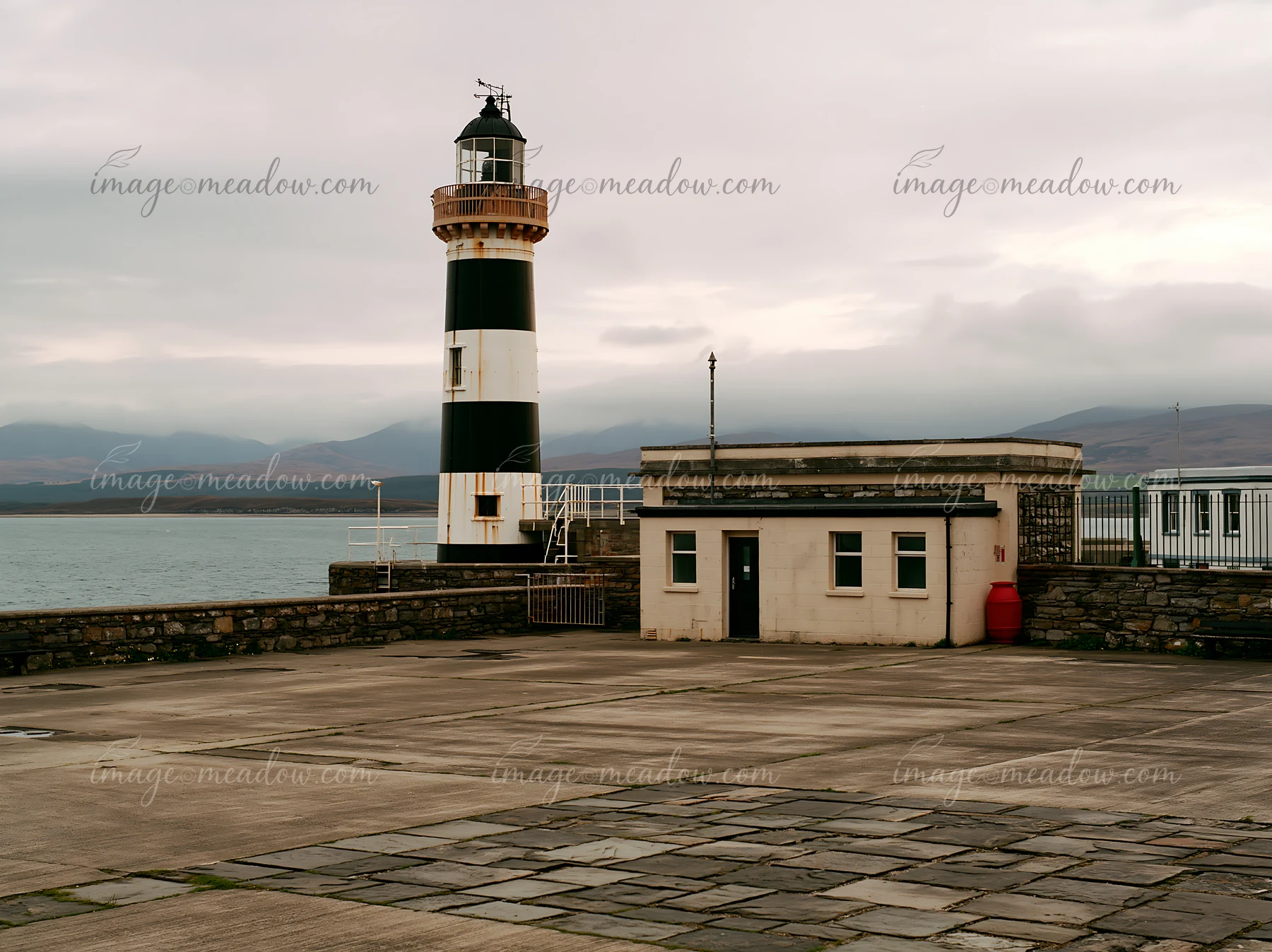 A lighthouse with black and white stripes stands near a simple coastal building, surrounded by calm waters and cloudy skies.
