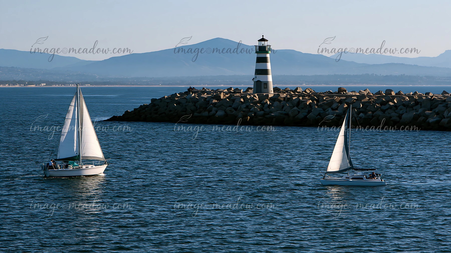 A lighthouse with black and white stripes stands on a rocky jetty as sailboats glide through the water, with mountains visible in the background during clear we
