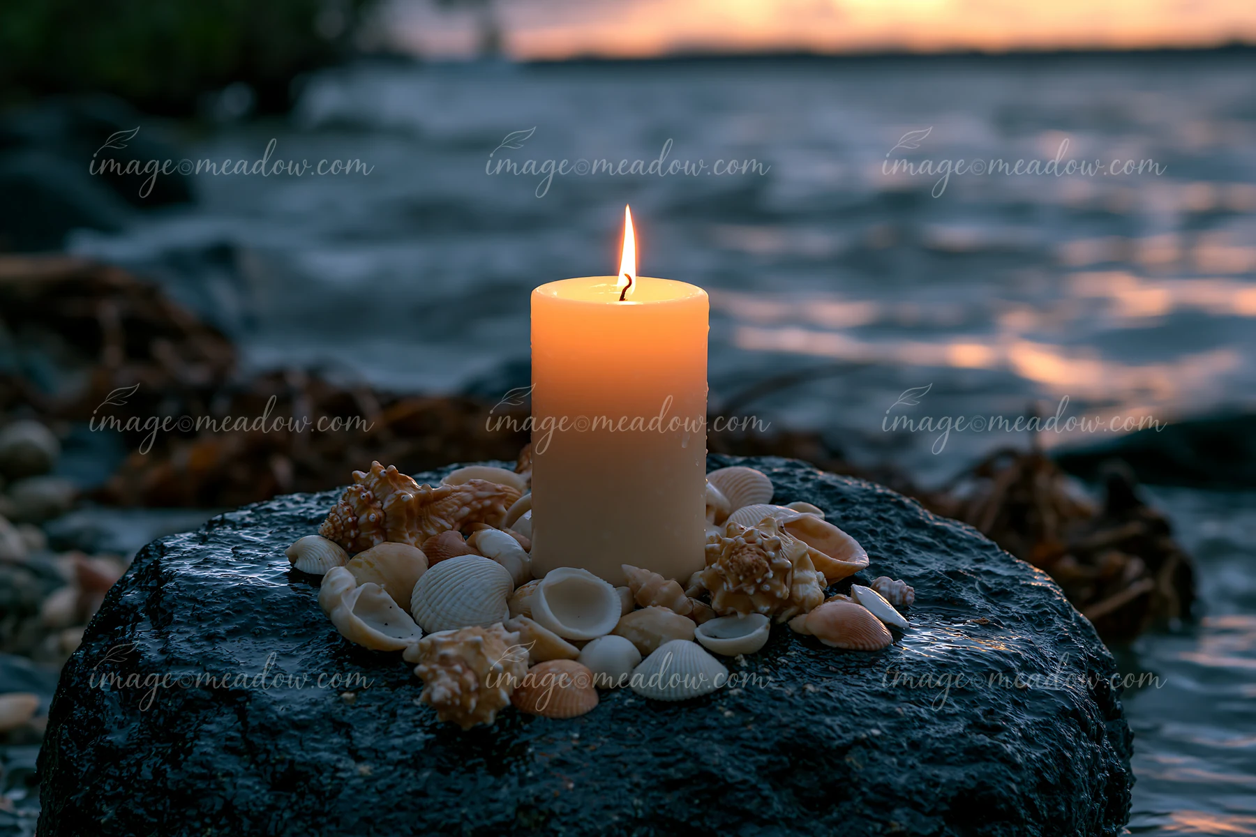 A lit candle stands on a dark rock surrounded by various seashells at the water's edge, capturing the warm light against a backdrop of rippling waves during twi