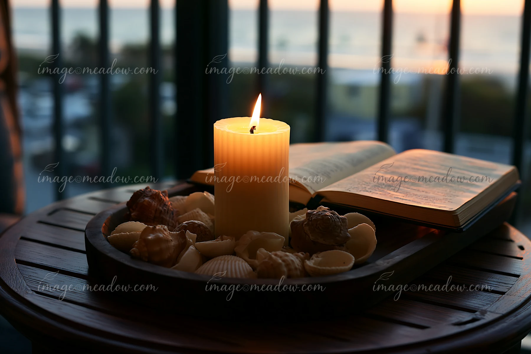 A lit candle stands on a tray filled with seashells next to an open book.