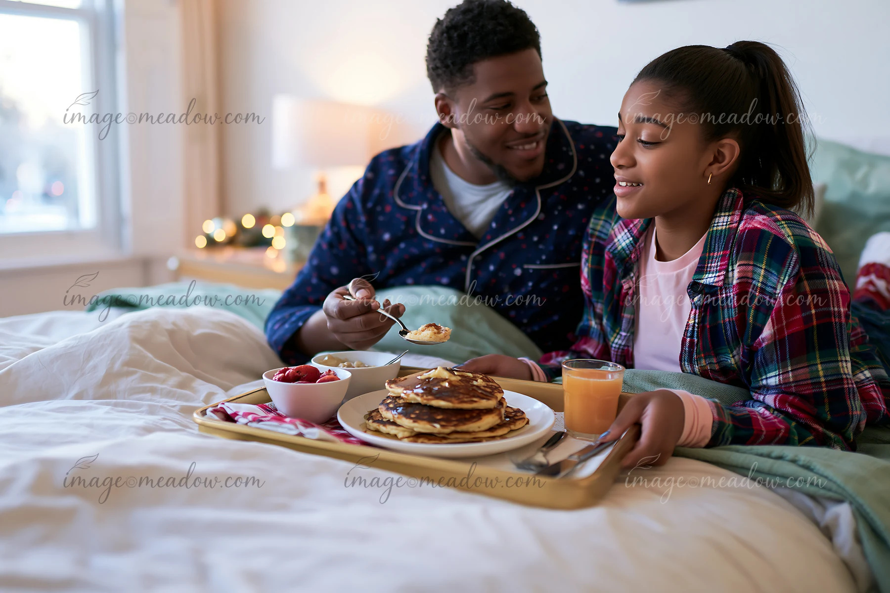 A man and a girl enjoy a breakfast tray in bed featuring pancakes, fruit, and juice.