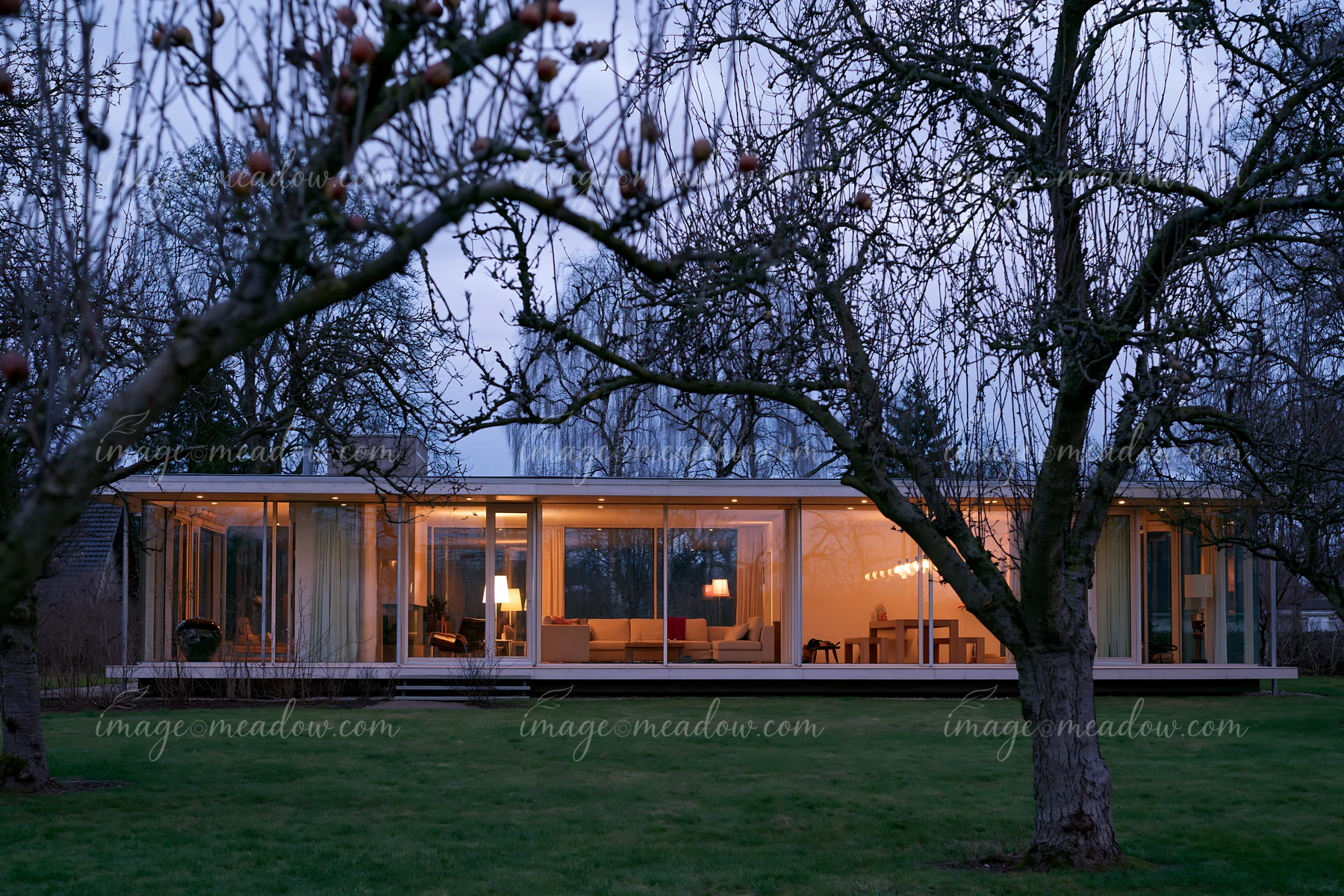 A modern empty garden house with glowing glass walls surrounded by trees as twilight approaches.