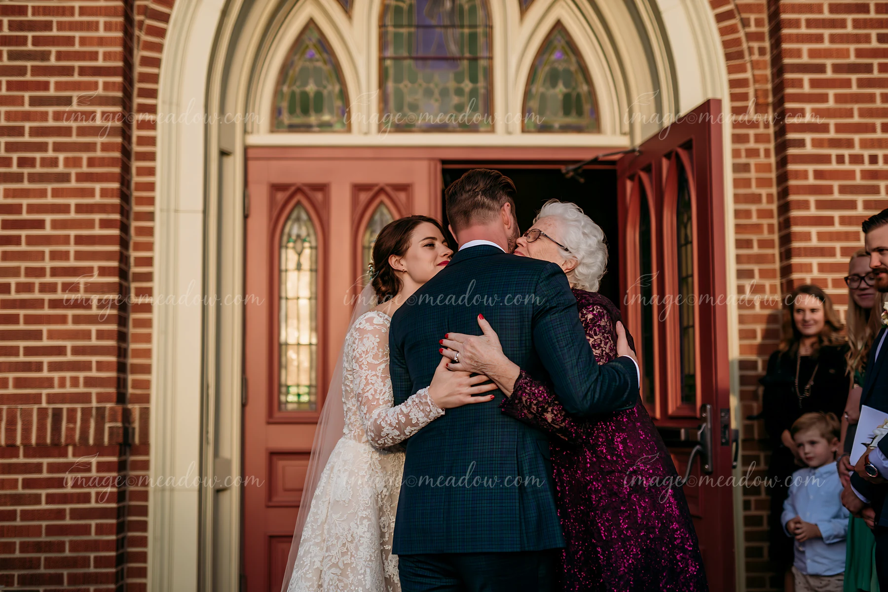 A newlywed couple stands at a church doorway, embracing relatives with joy.