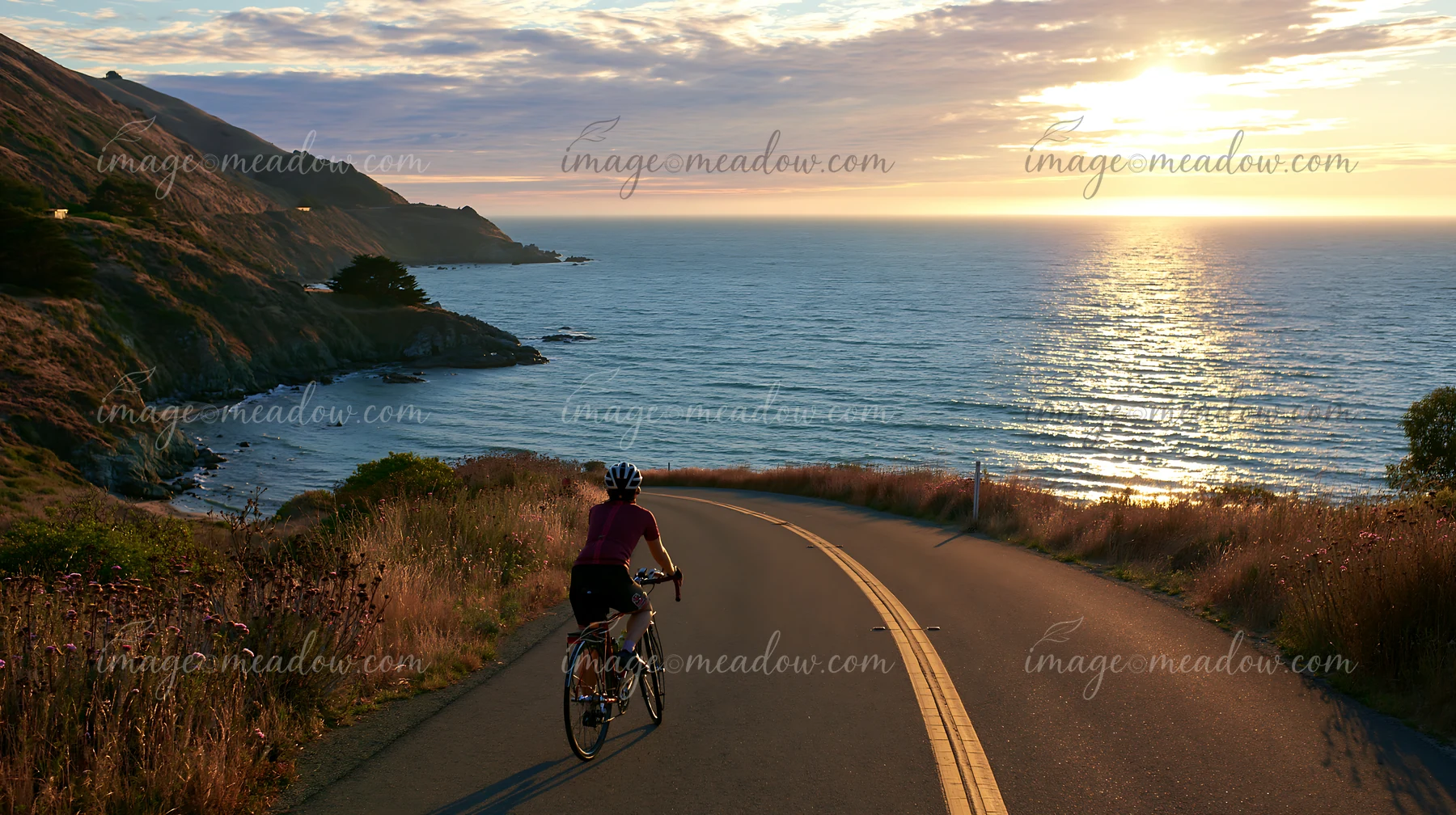 A person cycles along a road by the coastline with sheltered bay during sunset.