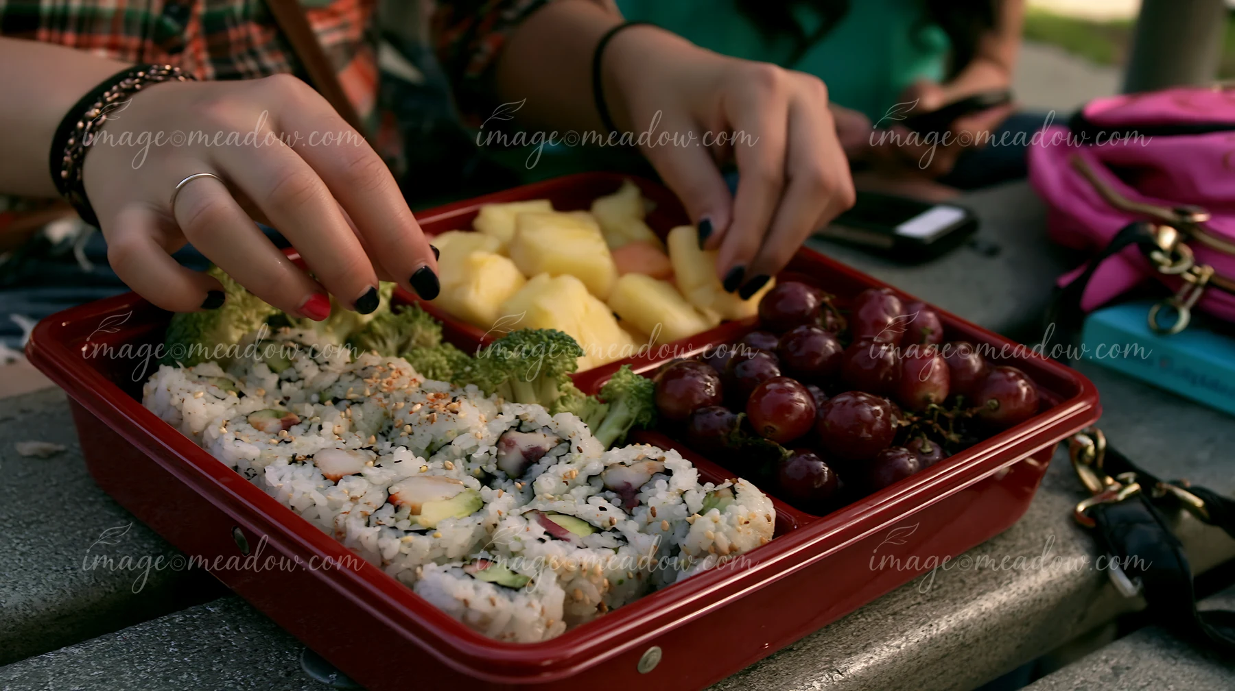A person enjoys a sushi lunch box with compartments at a picnic table.
