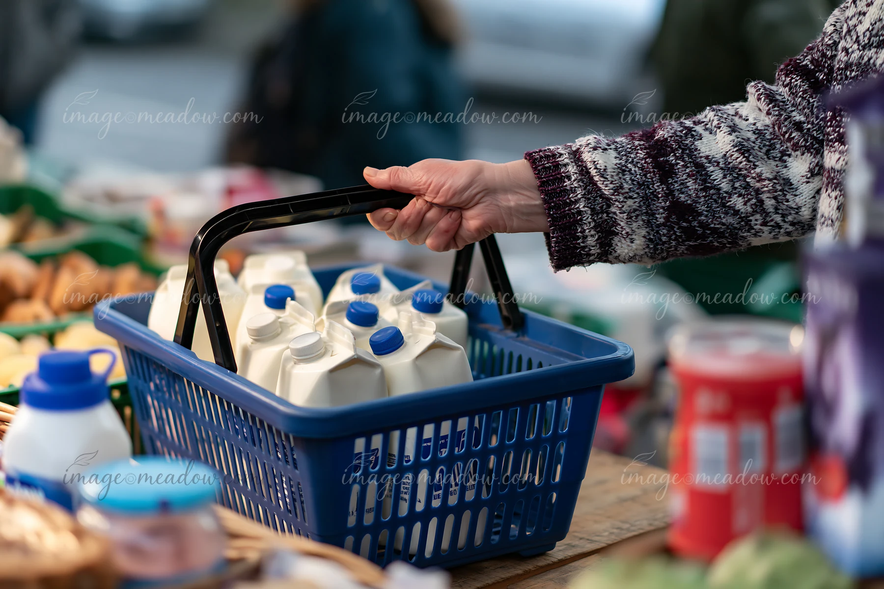 A person holds a blue shopping basket that contains milk cartons while standing at a market.