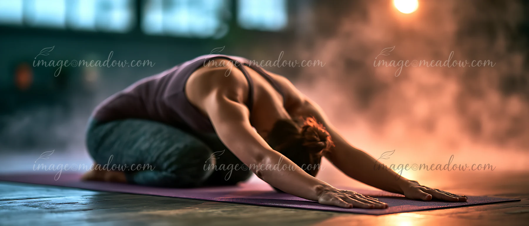 A person is performing child's pose on a yoga mat in a dimly lit studio filled with soft lighting and light mist.