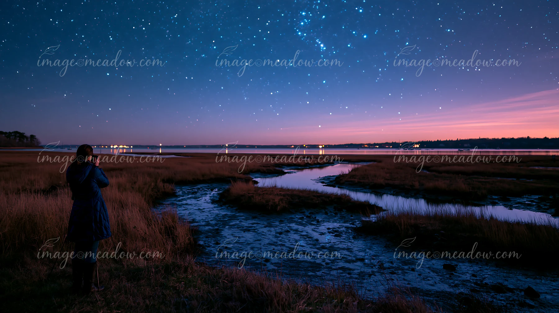 A person is photographing stars at night near a river with grassy banks.