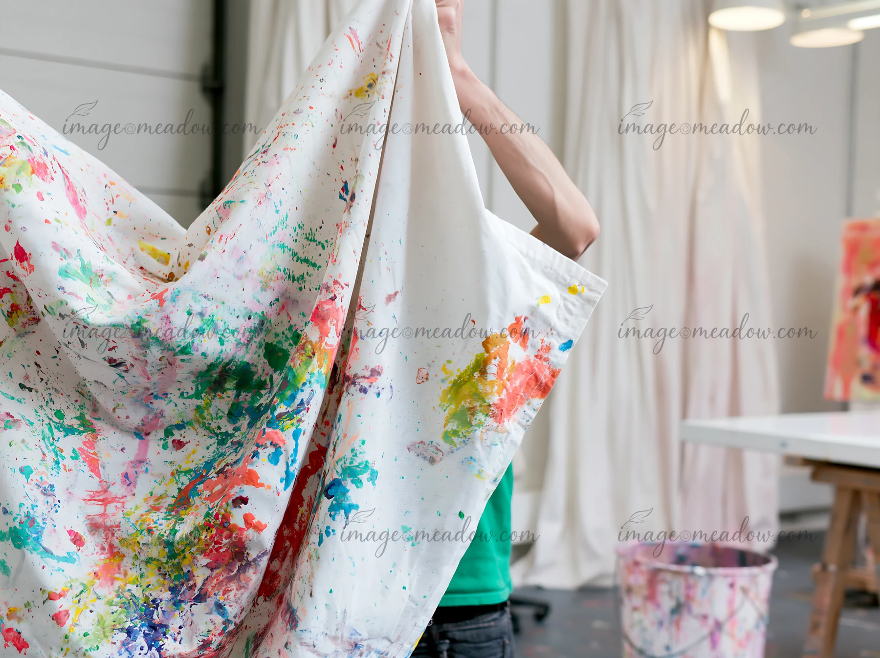A person lifts a piece of cotton fabric covered with colorful paint splatter in an art studio.