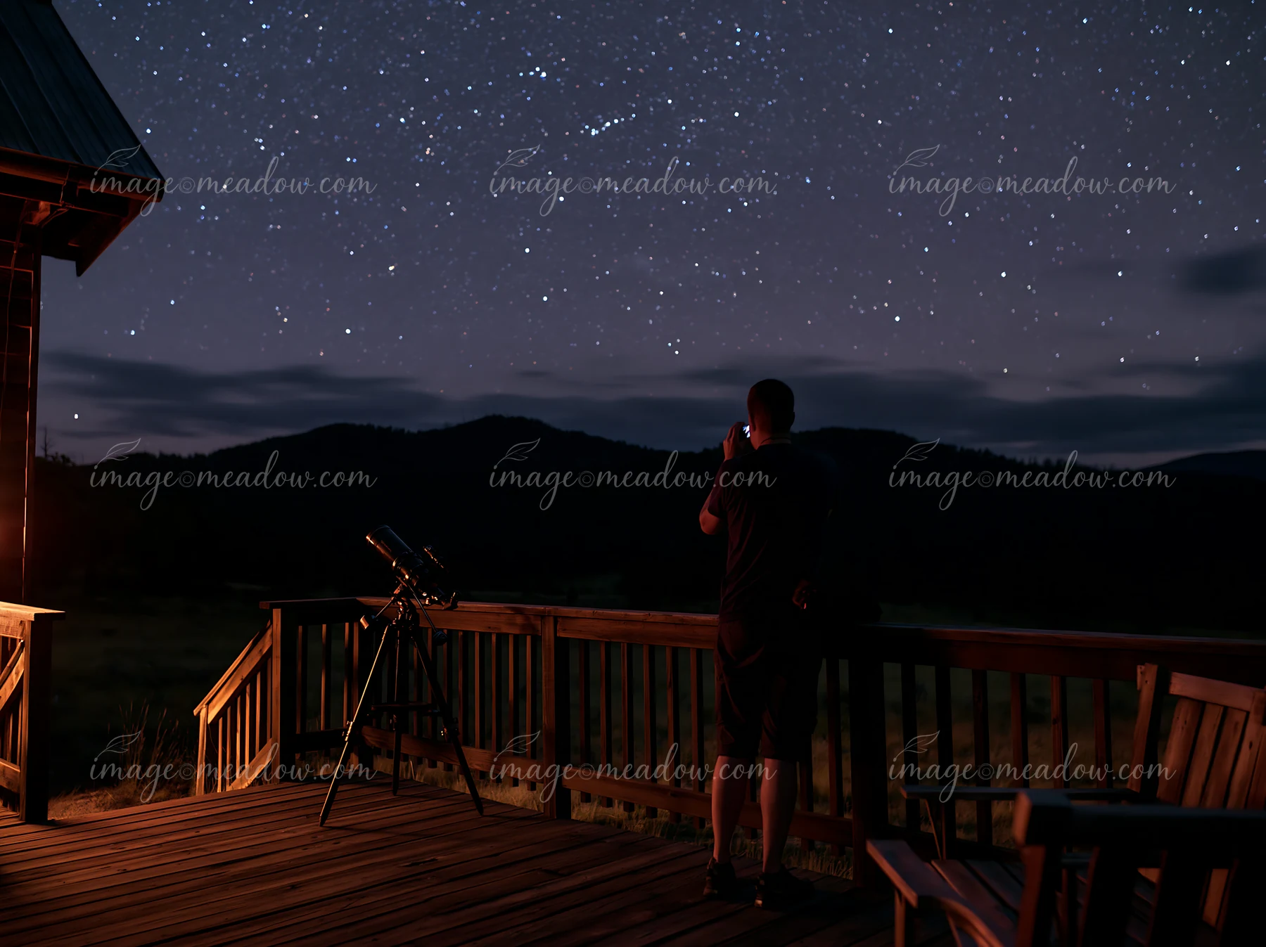 A person photographing stars from a mountain balcony with a telescope nearby.