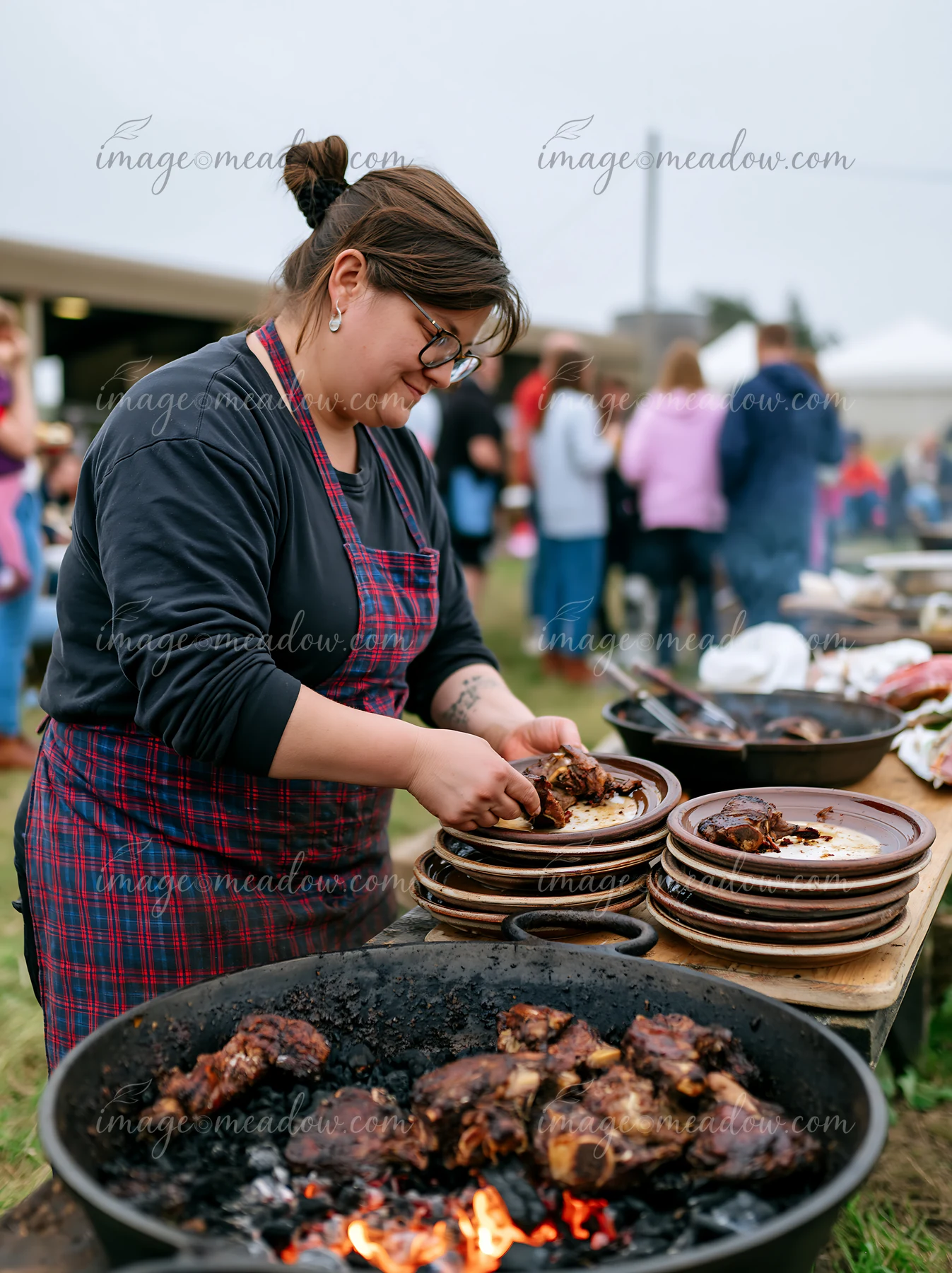 A person prepares goat meat on a grill while serving plates during a countryside festival.