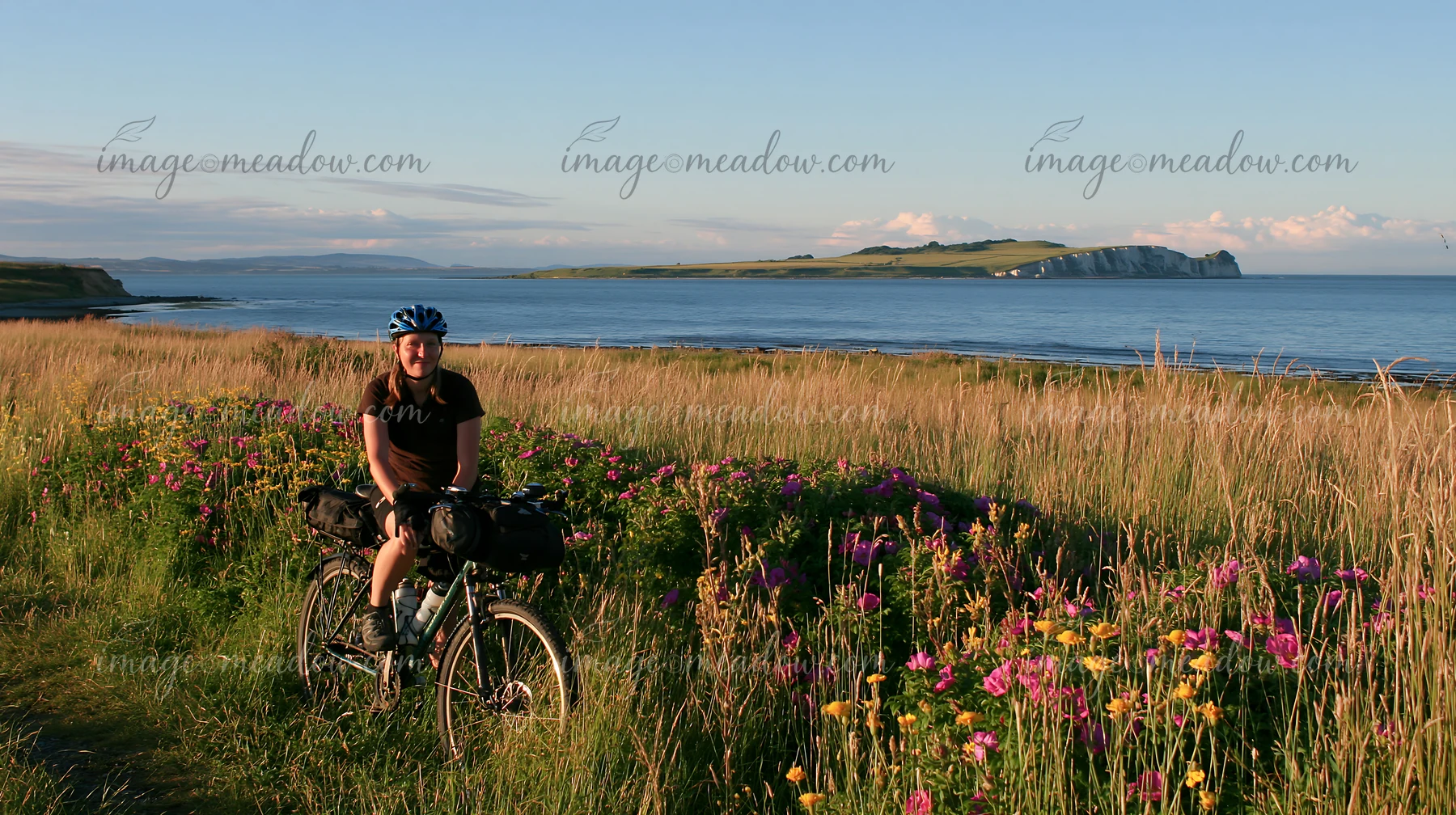 A person rides a bicycle along a path lined with flowers and tall grass, overlooking the ocean and white cliffs on an island in the distance during daylight.