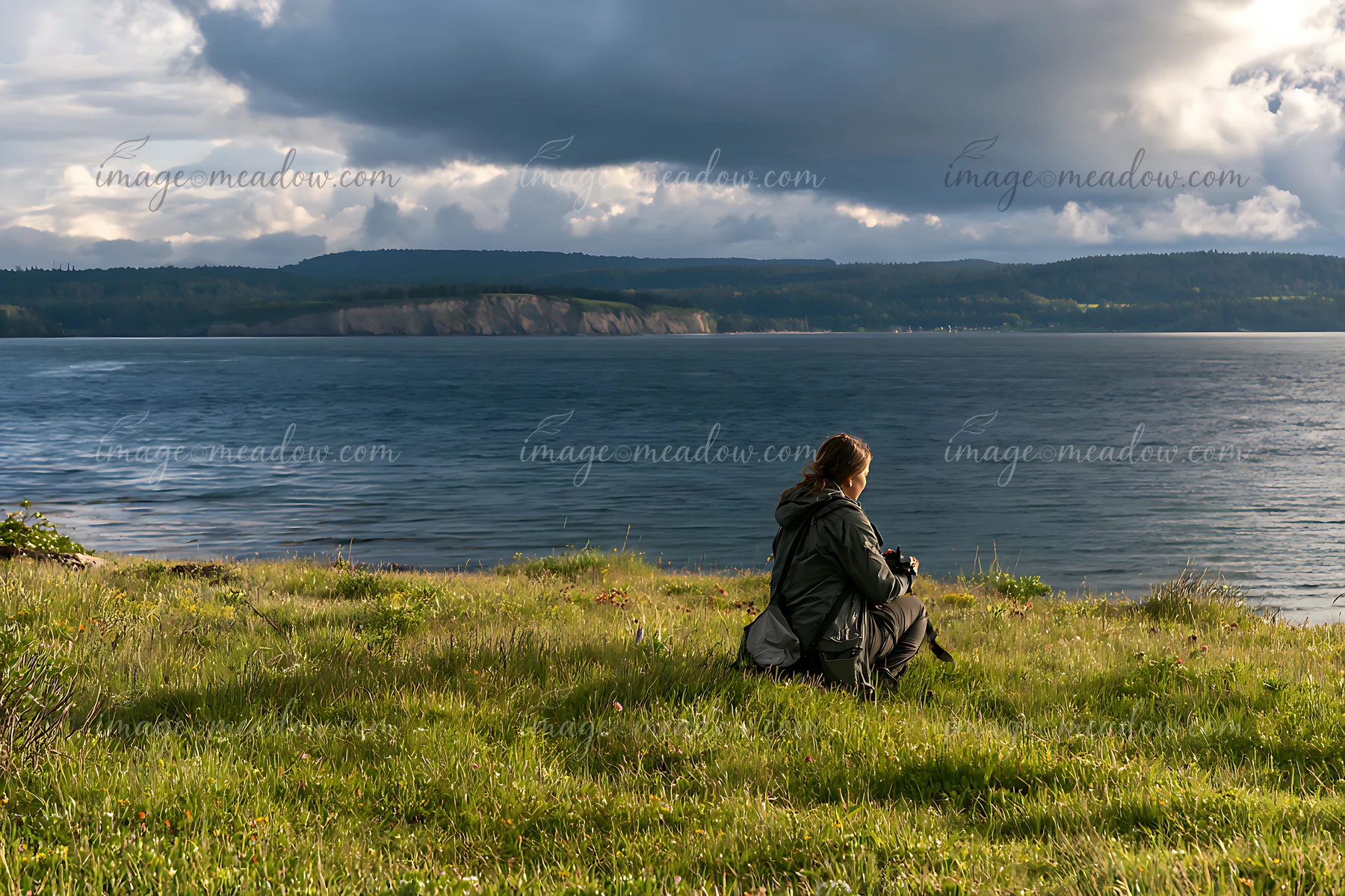 A person sits on a grassy area overlooking a body of water with dark clouds in the sky.